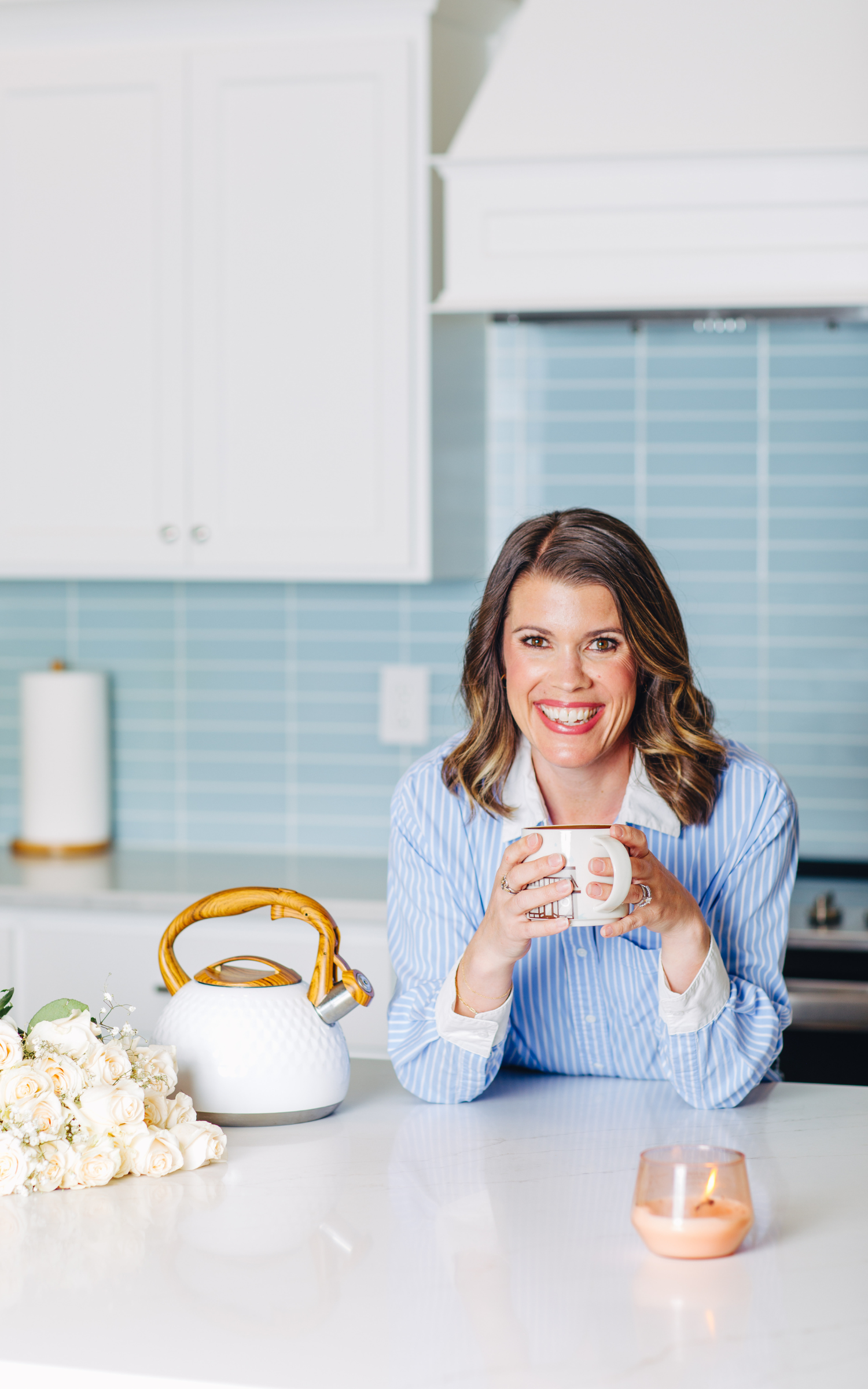 Woman with brown hair smiling and holding a white mug in a modern, bright kitchen with white cabinets, blue backsplash, and a bouquet of white roses on the counter.