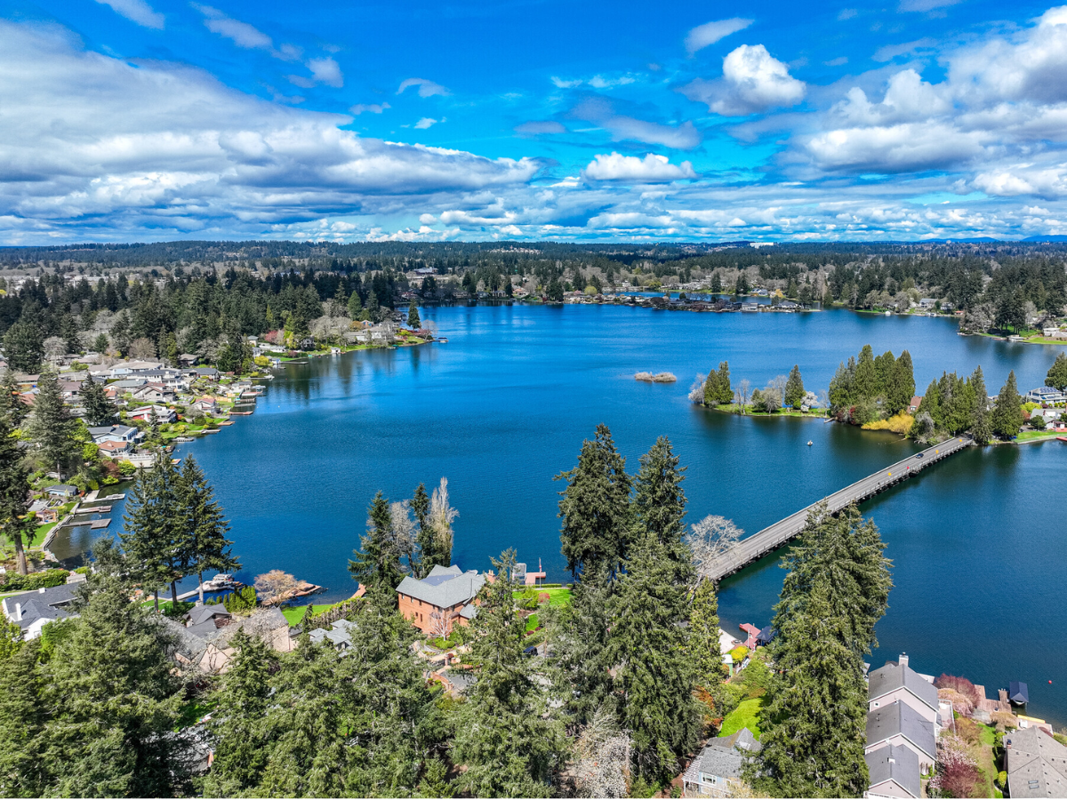 Aerial view of a lake with surrounding houses, trees, and a bridge crossing the water under a partly cloudy sky.