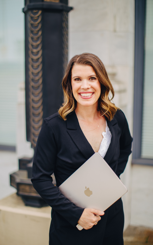 A woman with shoulder-length brown hair smiling and holding a closed MacBook laptop. She is standing outdoors in front of a light-colored building with a window, wearing a black blazer and white blouse.