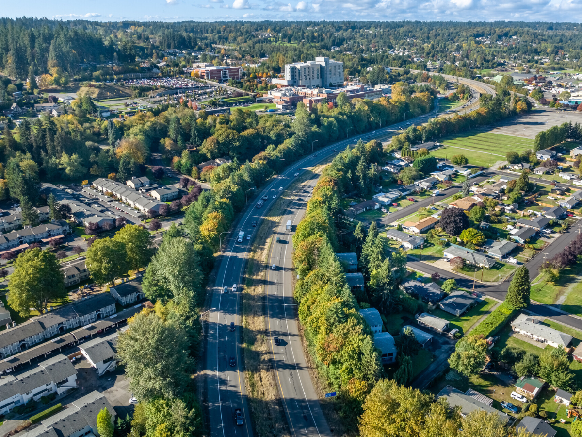 An aerial view of a suburban area with a curved highway, residential houses, and a large institutional building complex surrounded by trees and green spaces, with distant hills and a partly cloudy sky.