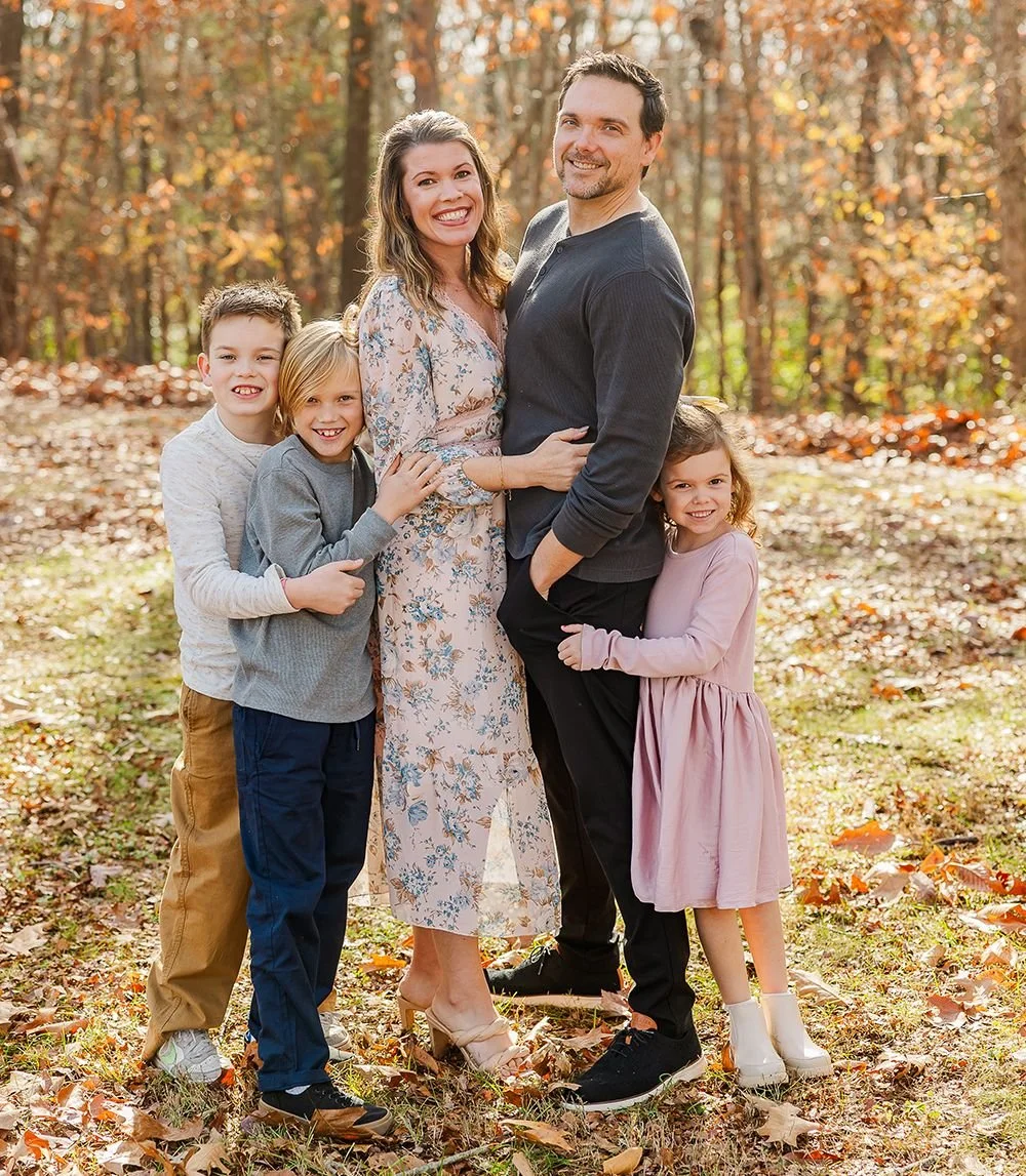 A family of six standing outdoors in a wooded area during fall, smiling at the camera. The family includes two adults and four children, with autumn leaves scattered on the ground.