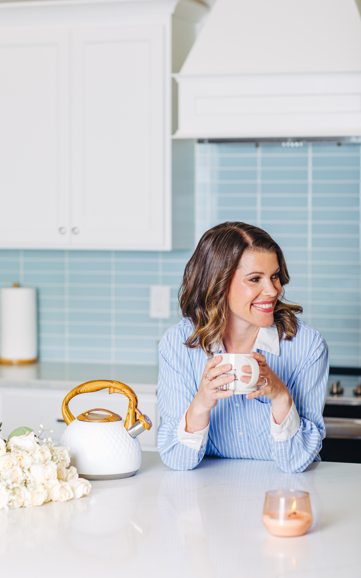 A woman with shoulder-length brown hair wearing a blue and white striped shirt is sitting at a white kitchen counter, holding a white mug and smiling. There is a bouquet of white roses, a white teapot with a wooden handle, and a lit candle on the counter. The background features light blue subway tiles, white cabinets, and a paper towel roll.