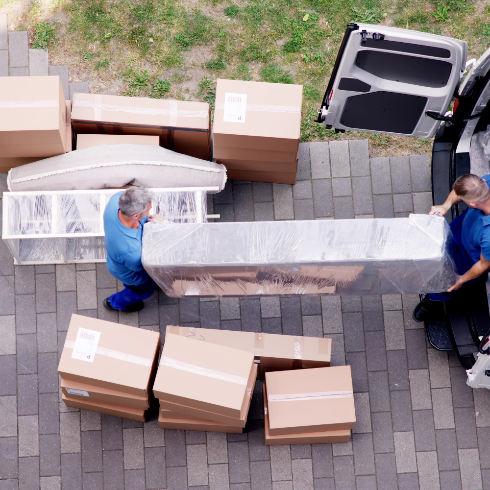 Two workers in blue scrubs move a wrapped couch into a delivery van. Several cardboard boxes and large packed items are on the ground and in front of the van on a paved surface.
