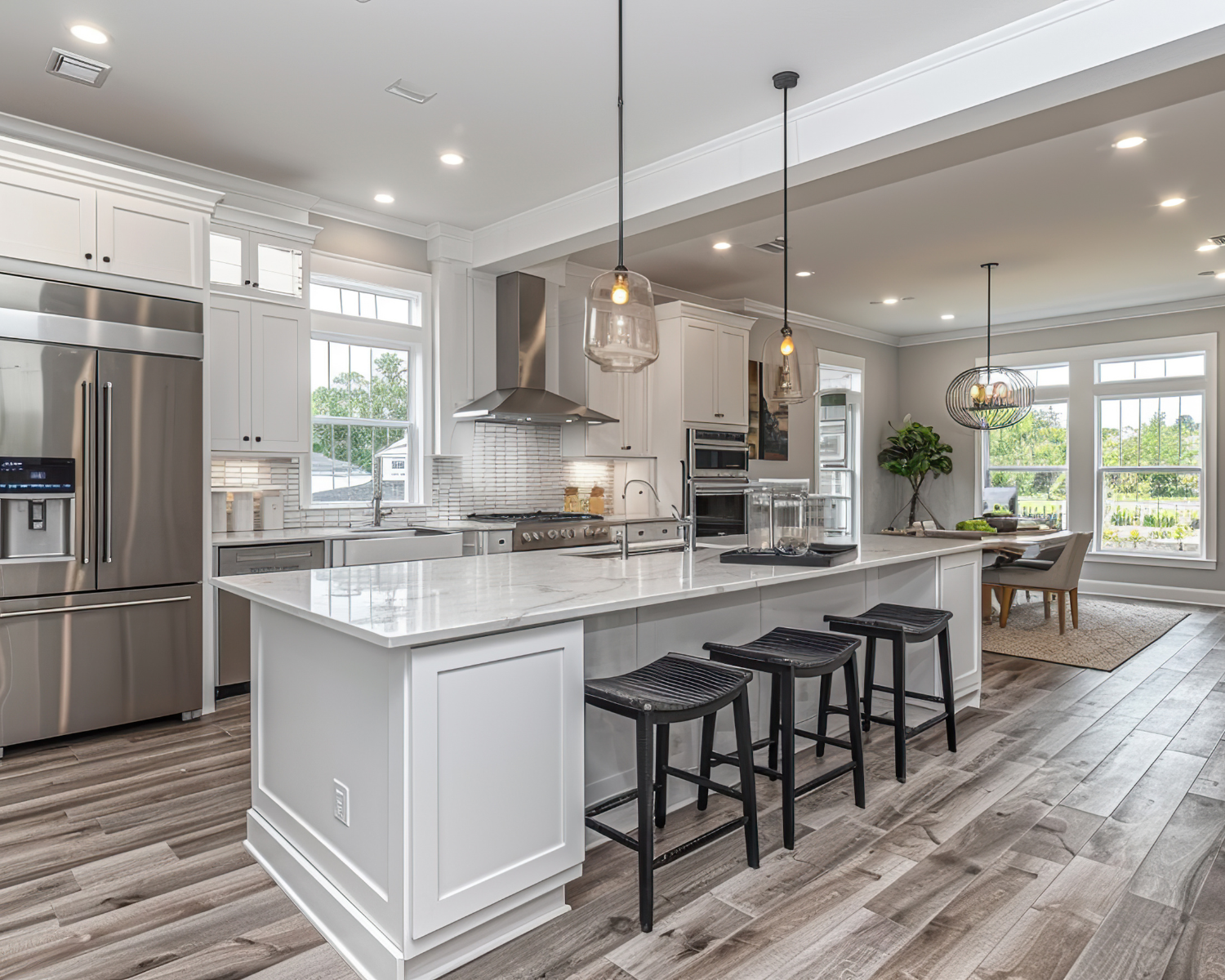Modern white kitchen with island, stainless steel appliances, pendant lighting, and wooden flooring, adjacent to a dining area with large windows and natural light.