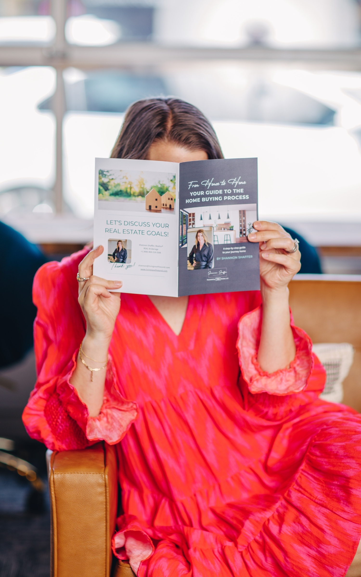 A woman with dark brown hair wearing a bright red, flowing dress with long sleeves, sitting on a tan chair in a well-lit indoor space, holding and reading a brochure about home buying.