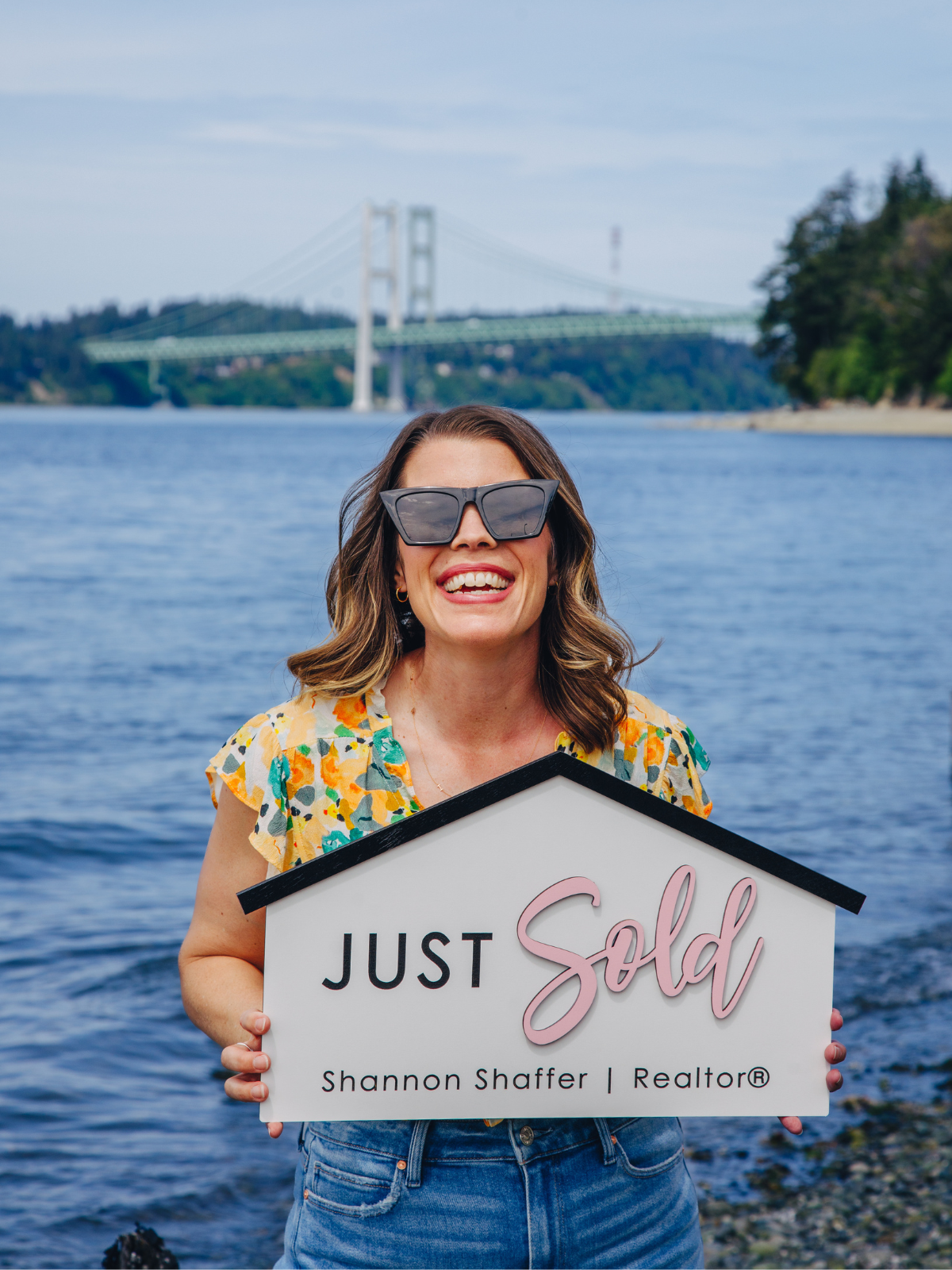 A woman with wavy brown hair wearing large sunglasses, a floral shirt, and jeans, smiling and holding a sign that says "Just Sold" with her realtor's name, Shannon Shaffer, in front of a body of water with a bridge in the background.