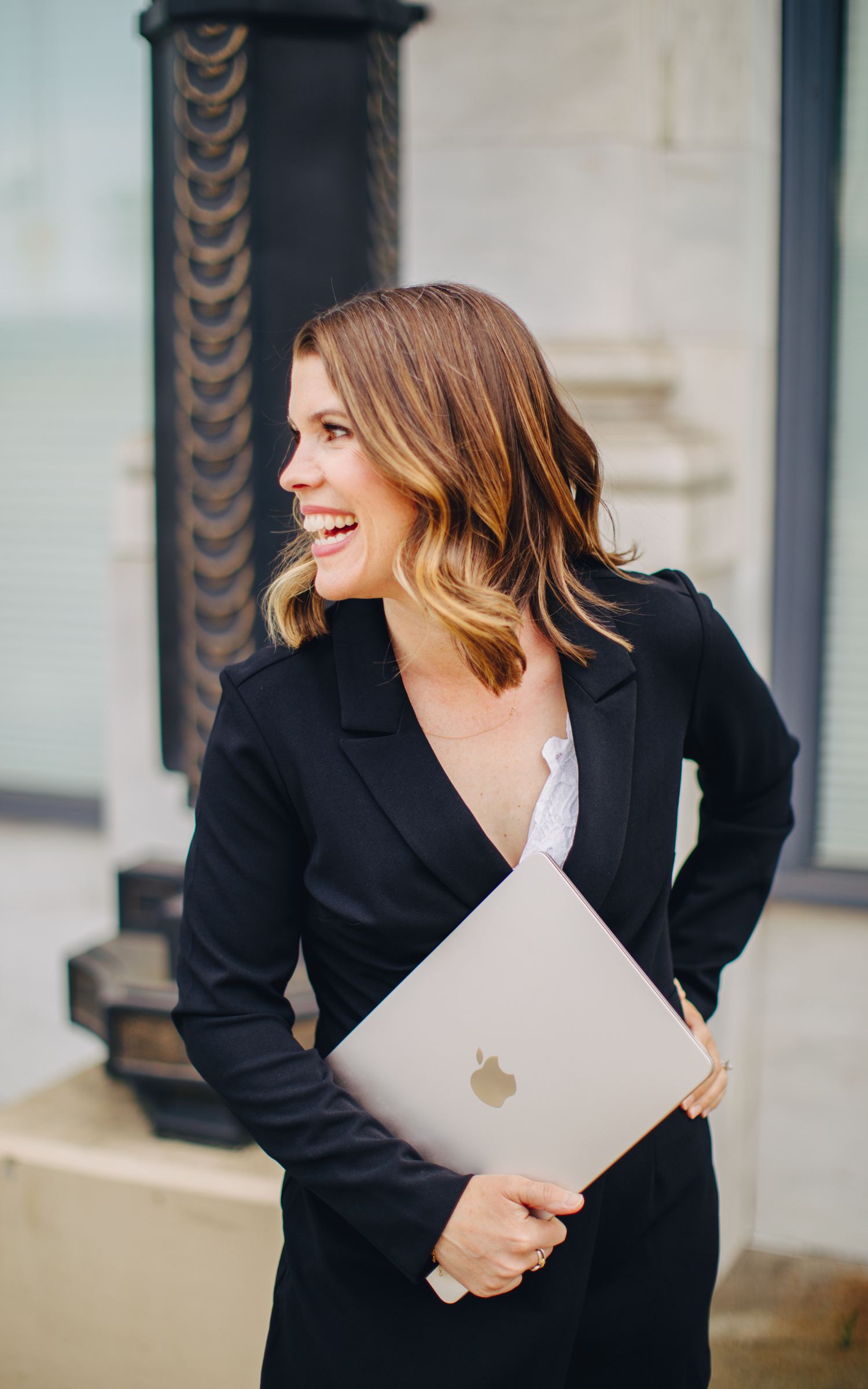 A woman dressed in a business suit holding a silver MacBook with an Apple logo, standing outdoors in front of a building with large windows.