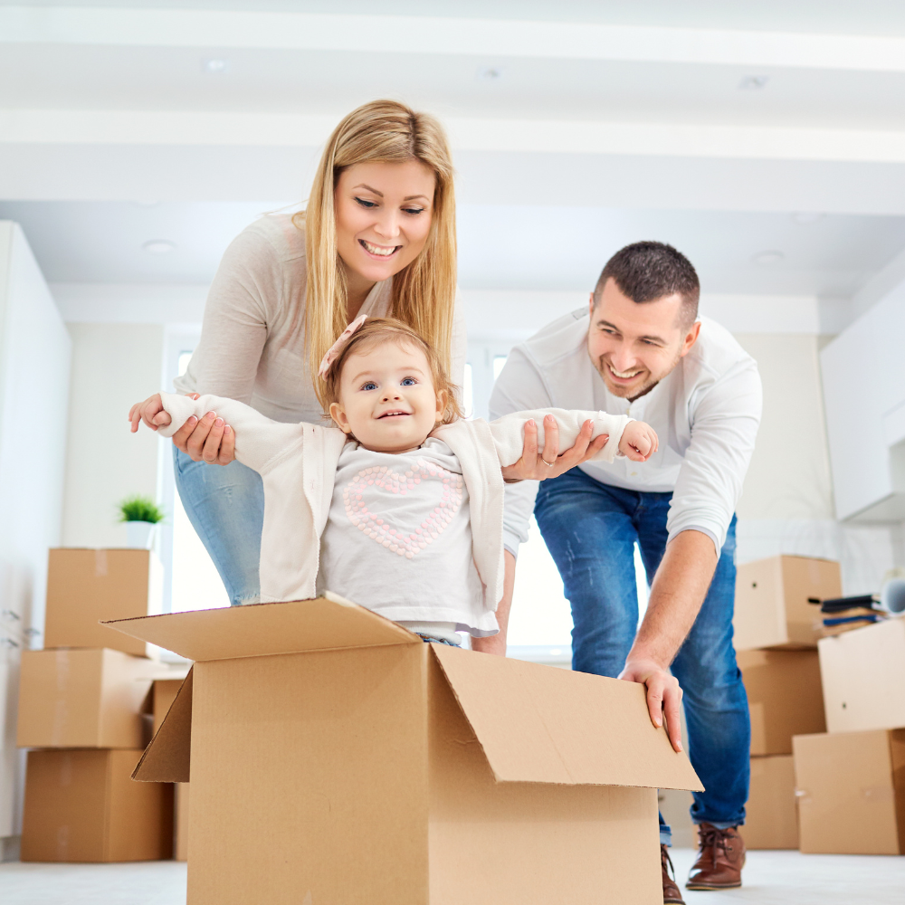 A family of three, a mother, father, and young daughter, joyfully unpacking in their new home, with the daughter standing in a cardboard box.