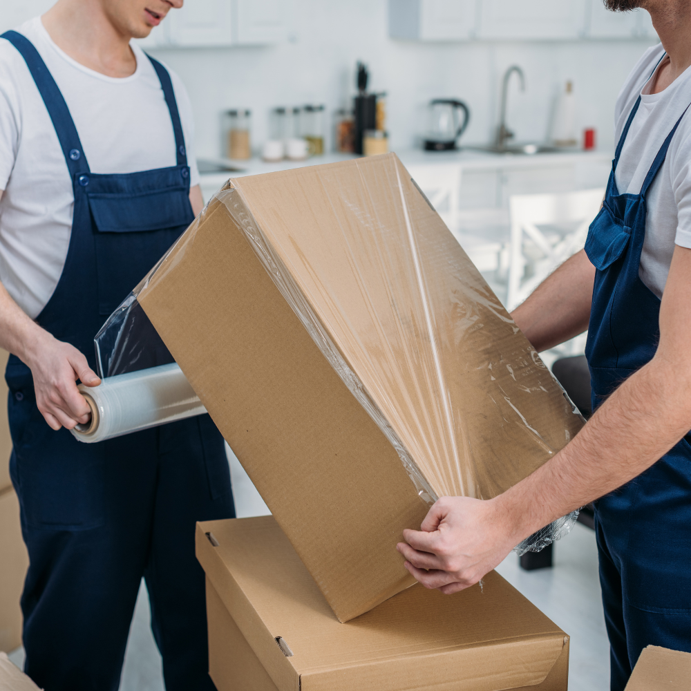 Two men in blue overalls packing a cardboard box with plastic wrap in a bright kitchen.