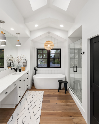 Bathroom with a white freestanding bathtub, black faucet, window, and walk-in shower with glass door, black hardware, black stool, wood flooring, white walls, and vaulted ceiling with skylights and hanging light fixture.