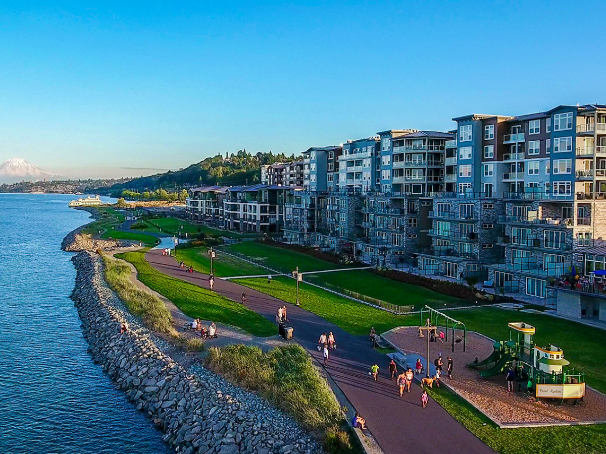 A scenic waterfront park with a walking path, green lawns, modern apartment buildings, and a playground, with a distant mountain in the background on a clear day.