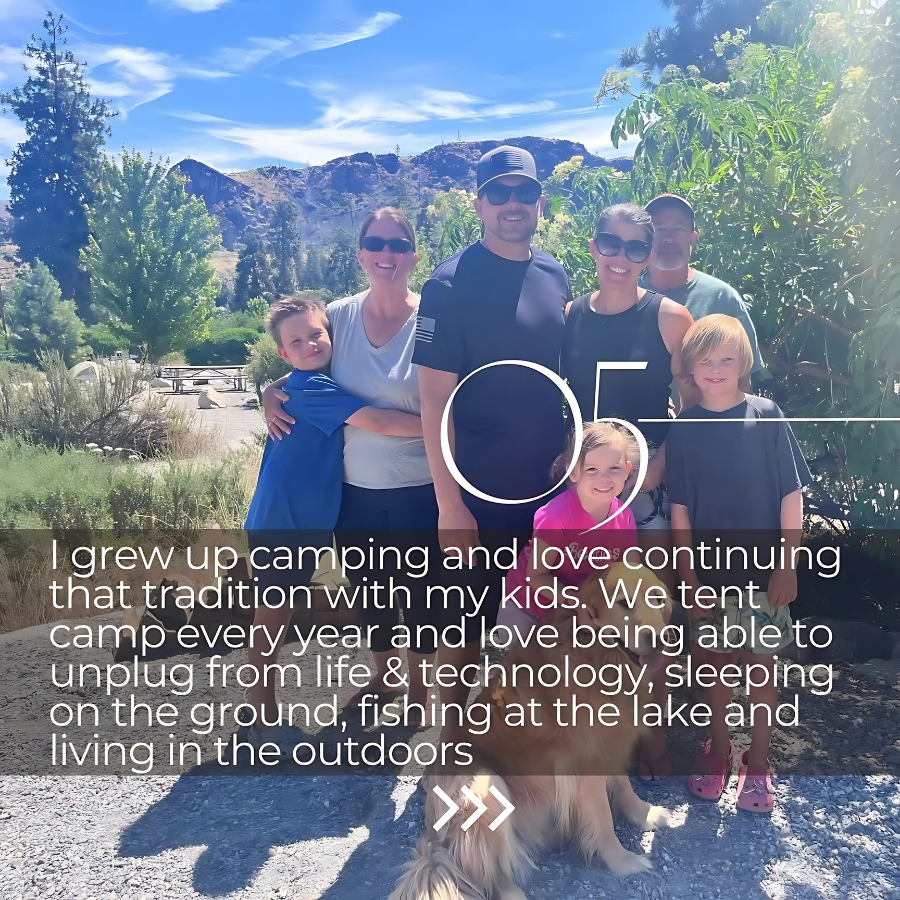 A group of seven people, including children and adults, posing outdoors in a scenic natural setting with mountains, trees, and blue sky in the background. They are smiling and appear to be enjoying a camping trip.