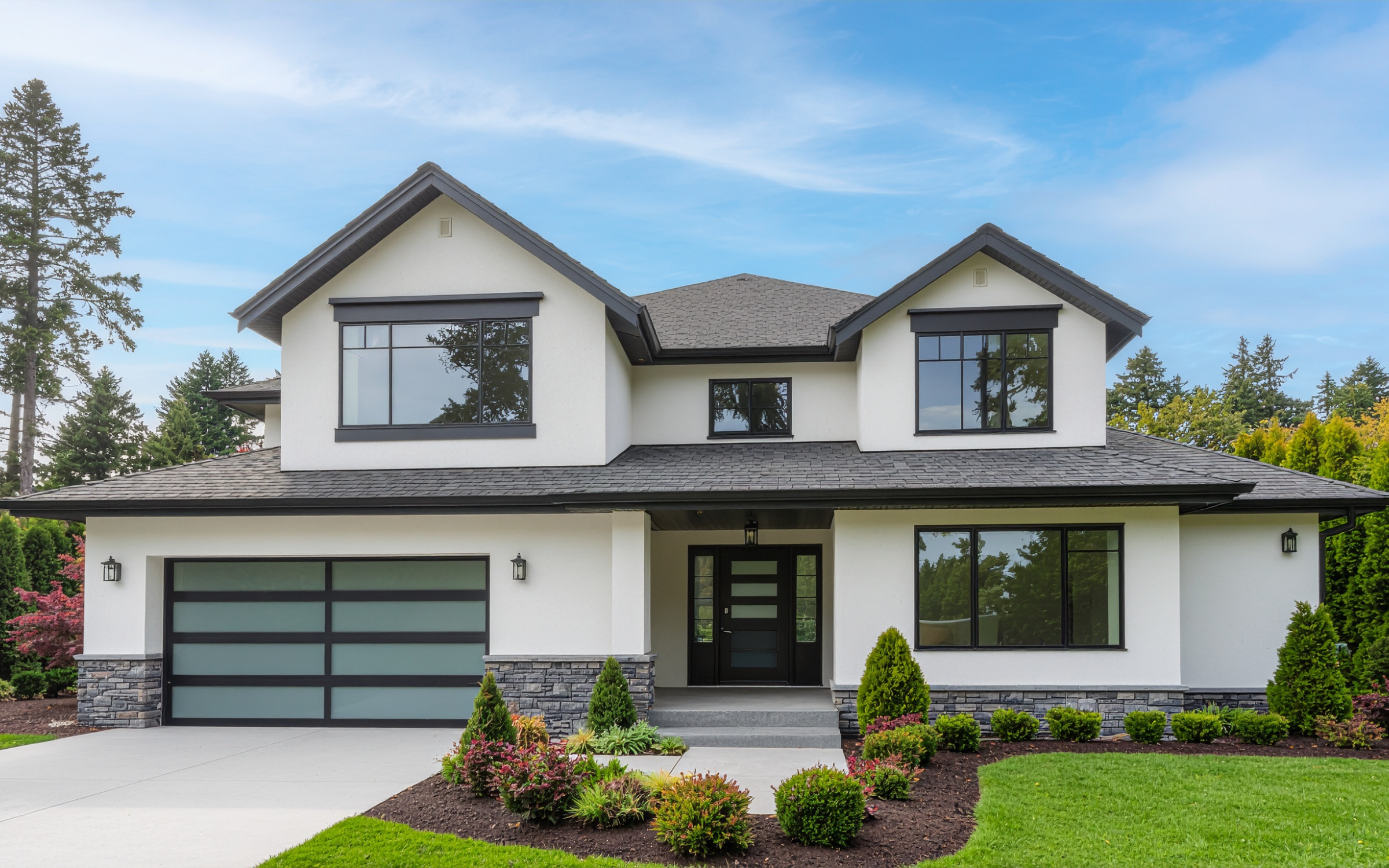 Modern two-story house with a white exterior, black accents, large windows, a stone foundation, a front yard with green grass and landscaped plants, and a driveway leading to a garage.