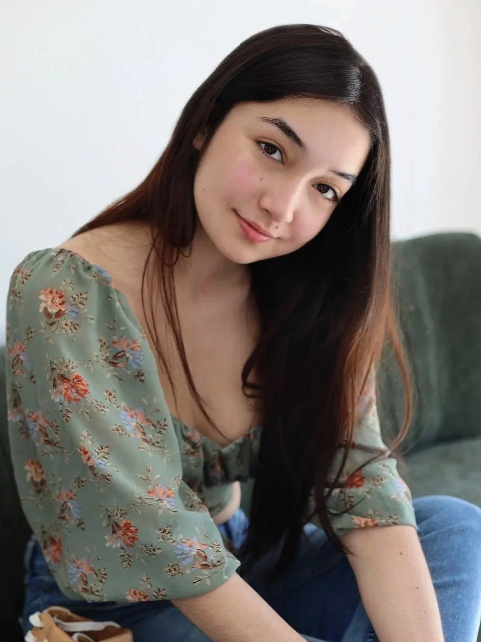 A young woman with long dark hair, wearing a light green floral blouse, sitting on a dark green couch, smiling at the camera.