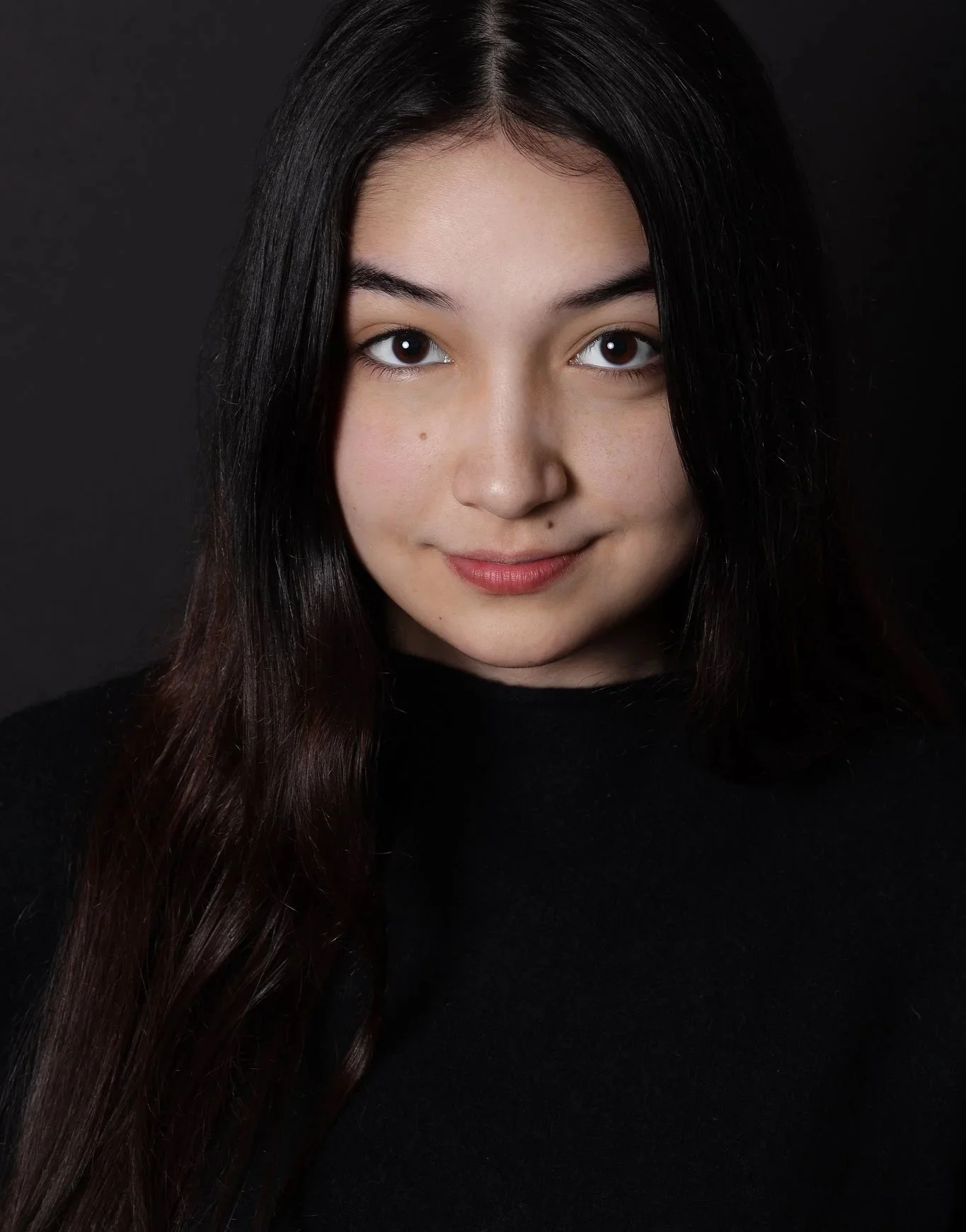 A young woman with long dark hair wearing a black top, looking at the camera against a dark background.