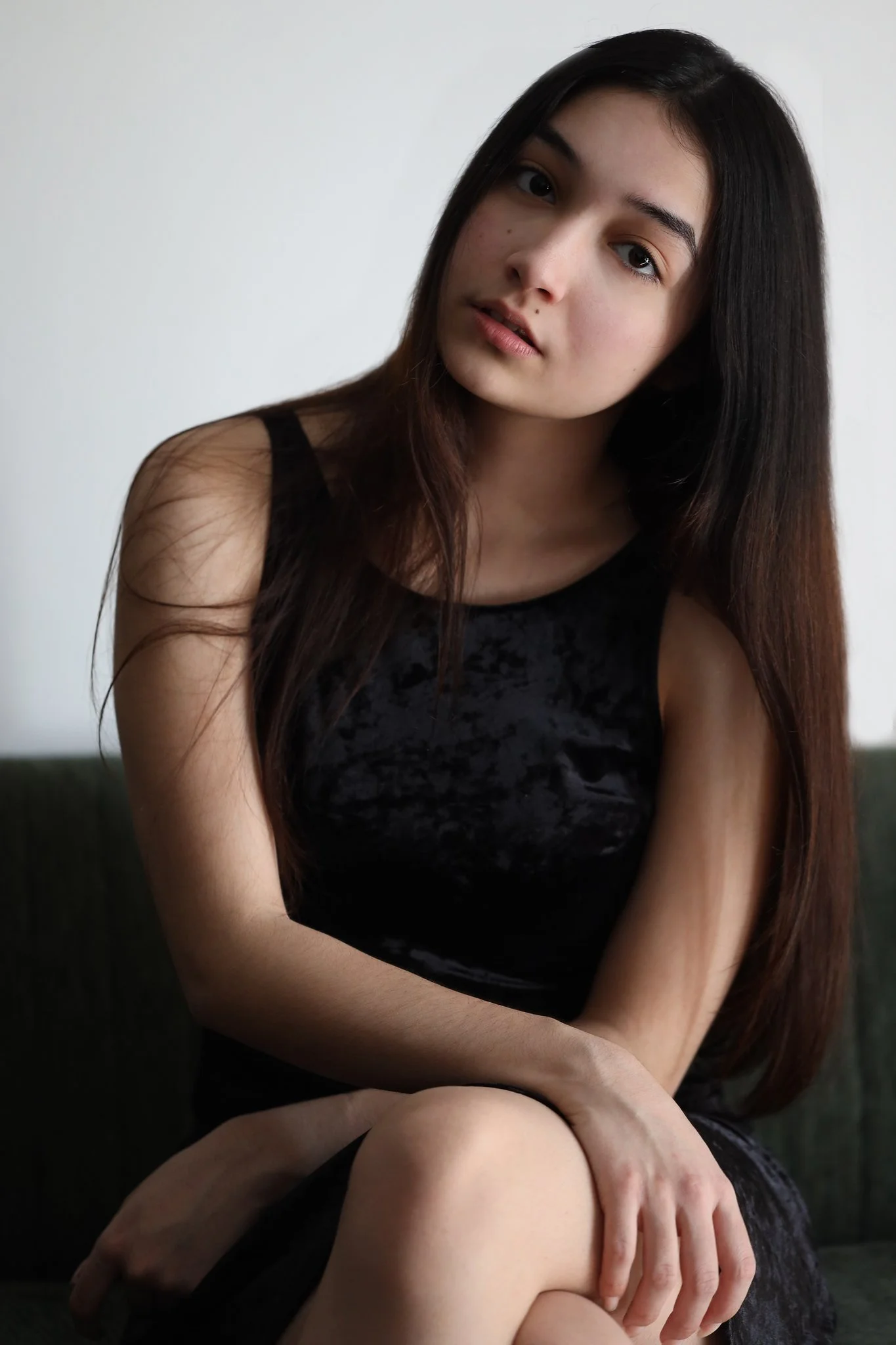 A young woman with long dark hair sitting on a dark green couch, wearing a black sleeveless dress, with her knees bent and her hand resting on her leg, looking at the camera.