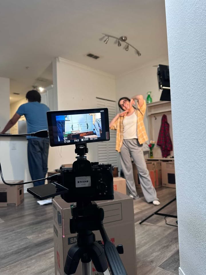 A woman wearing glasses, a yellow plaid shirt, and gray pants is posing for a camera in a room with boxes and decorative items, while a man in a blue shirt is in the background.