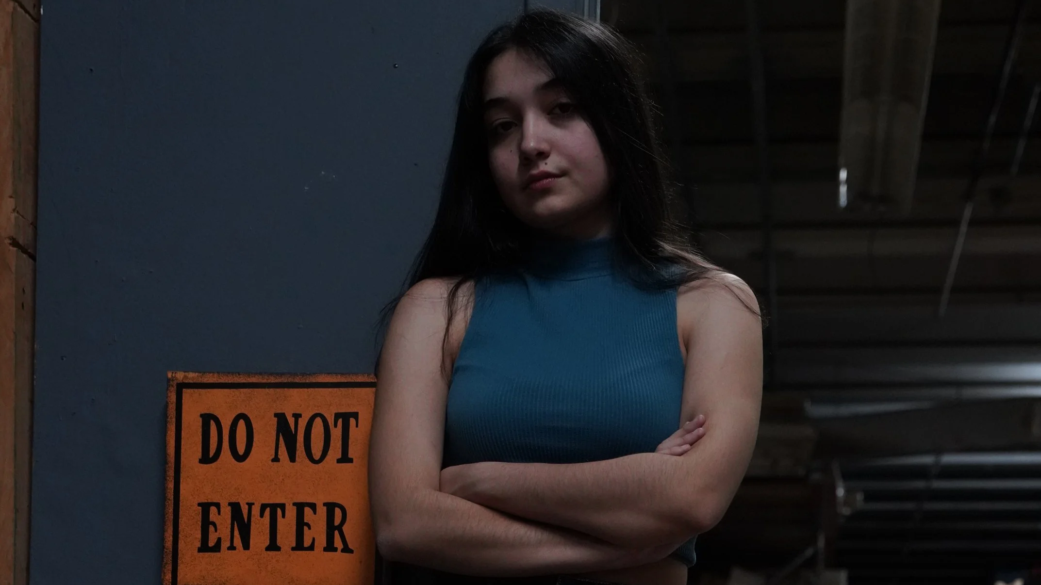 A young woman with long dark hair standing with crossed arms in front of a wall with an orange 'Do Not Enter' sign. She is wearing a sleeveless blue top and has a neutral expression.