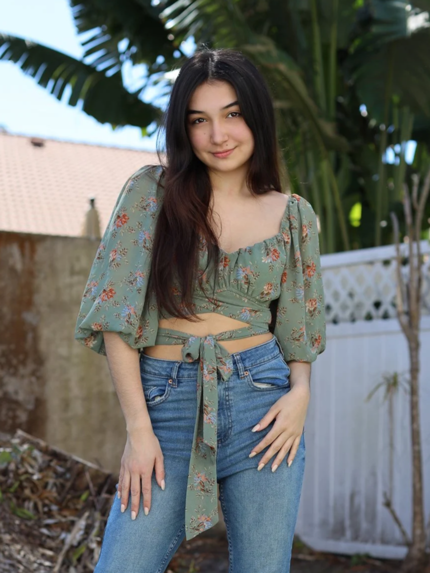 A young woman with long dark hair standing outdoors in front of a plastic white fence, wearing a green floral crop top with puffed sleeves and tied at the waist, paired with blue jeans. She is smiling softly, with trees and a house roof in the background.