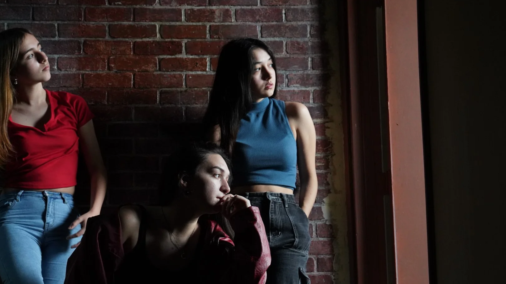 Three young women in casual clothing standing and sitting by a brick wall near a window, gazing outside thoughtfully.