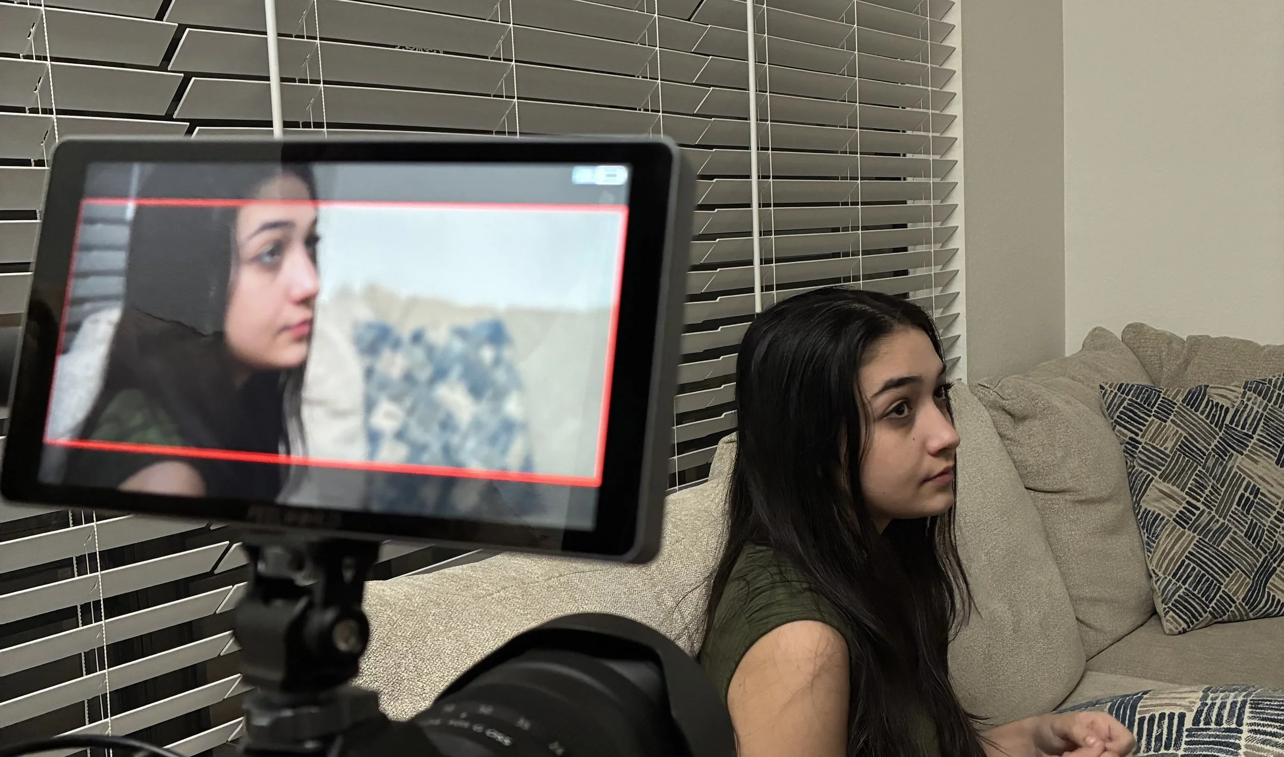 Woman sitting on a beige couch inside a living room with window blinds, with a camera capturing her profile.