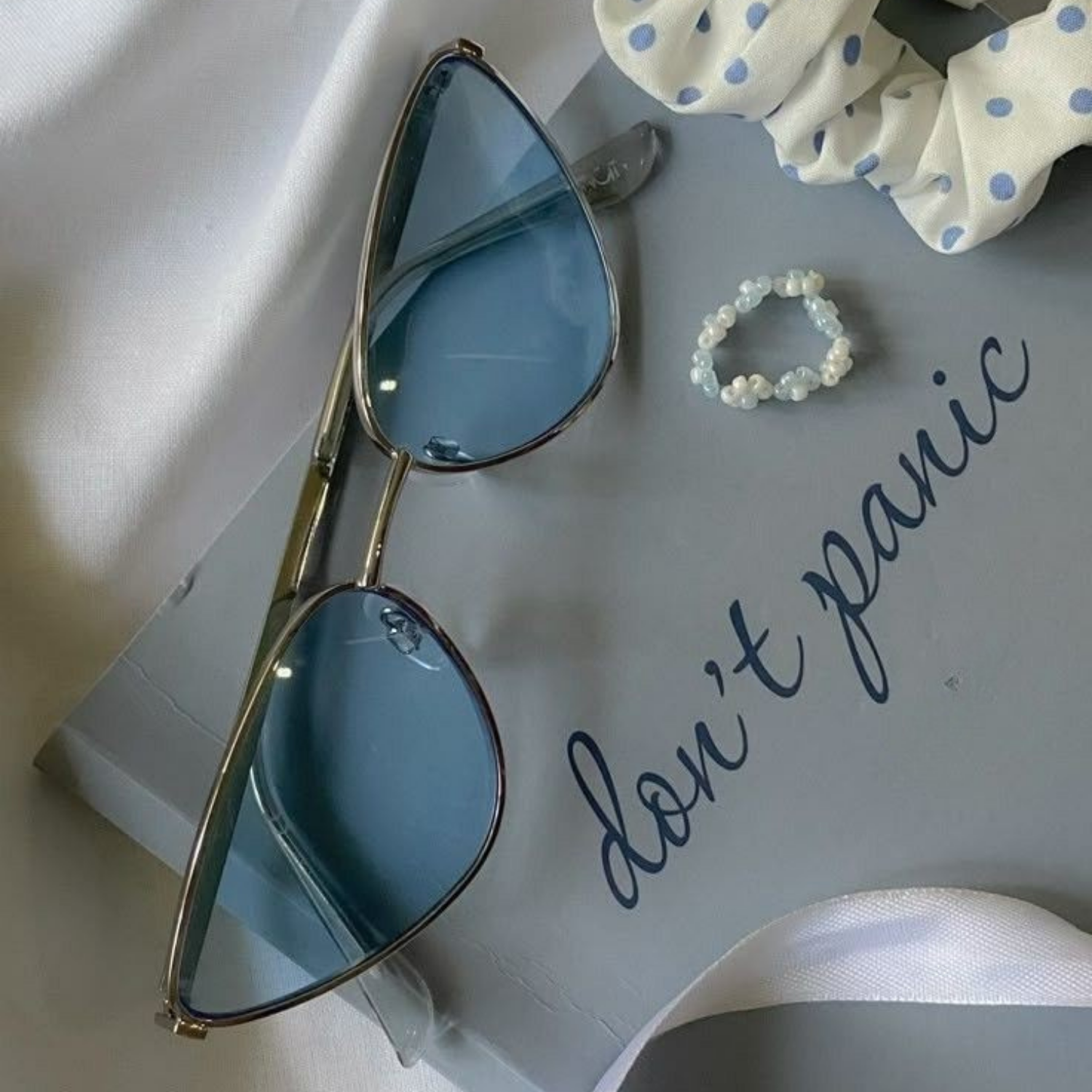 Blue-tinted sunglasses with gold frame placed on a white paper with handwritten text and next to a white cloth with blue polka dots, some of which is folded.