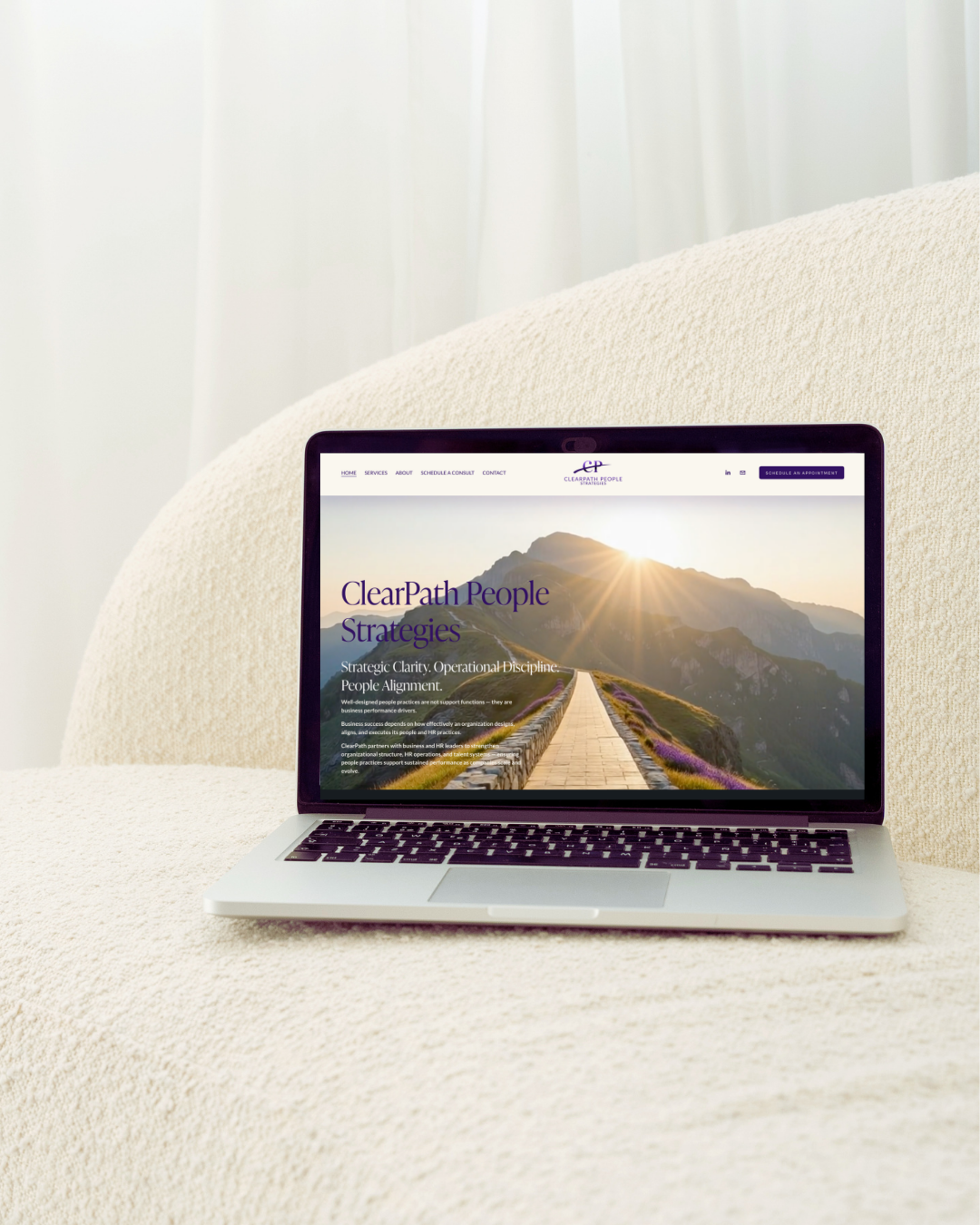 Laptop on a cream-colored textured couch displaying the homepage of a website called ClearPath People Strategies, featuring a mountain landscape with a sunrise.