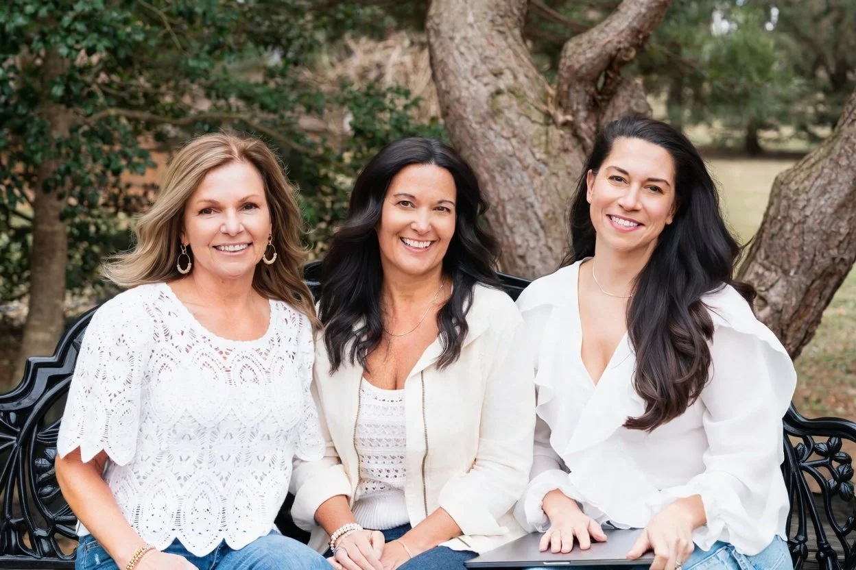 Three women sitting on a black bench outdoors with trees in the background, smiling at the camera.