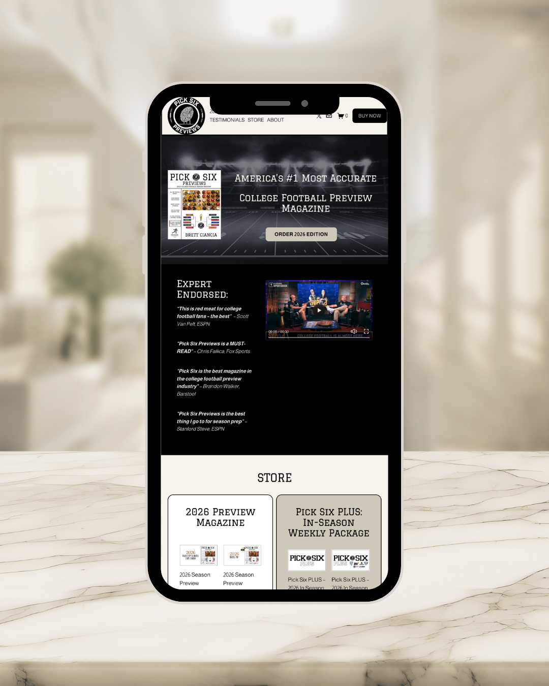 Mobile phone displaying a website for Pick Six Previews, a college football preview magazine, with sections about the 2026 edition and in-season weekly packages on a white marble surface with blurred indoor background.