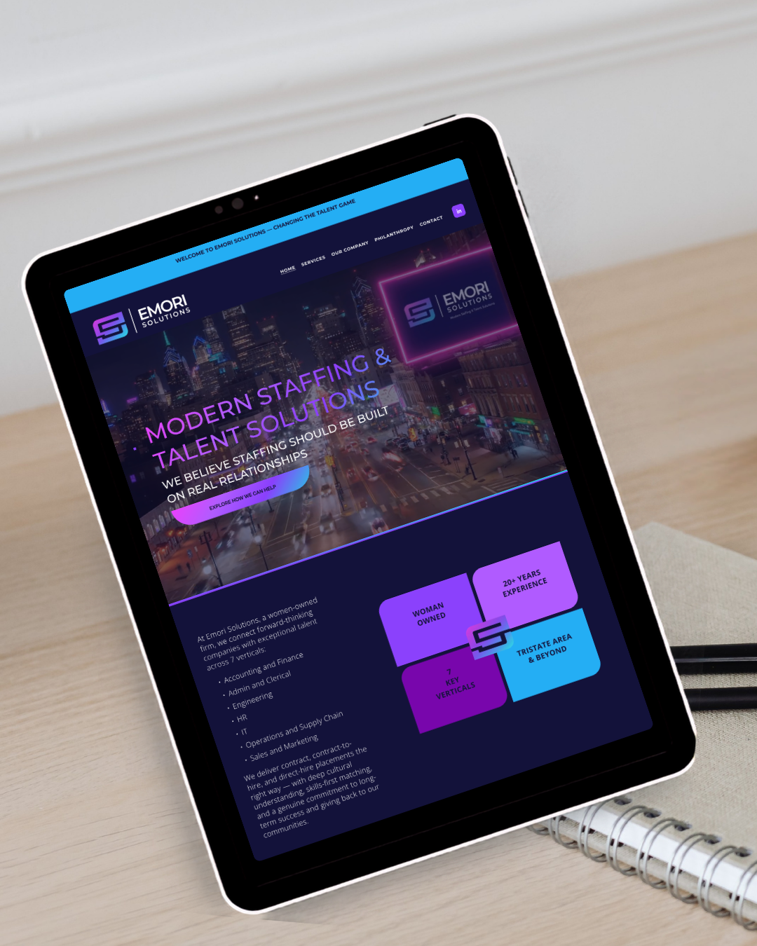 Tablet displaying the homepage of Emori Solutions, a staffing and talent solutions company, showing a cityscape background with neon graphics and text.