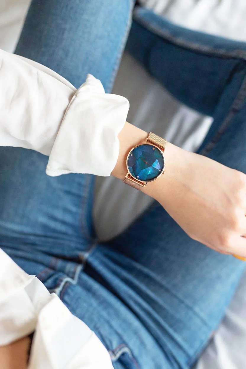 Close-up of a person's left wrist wearing a gold watch with a dark blue face, sitting on a blue denim chair, wearing a white long-sleeve shirt with the sleeve rolled up.