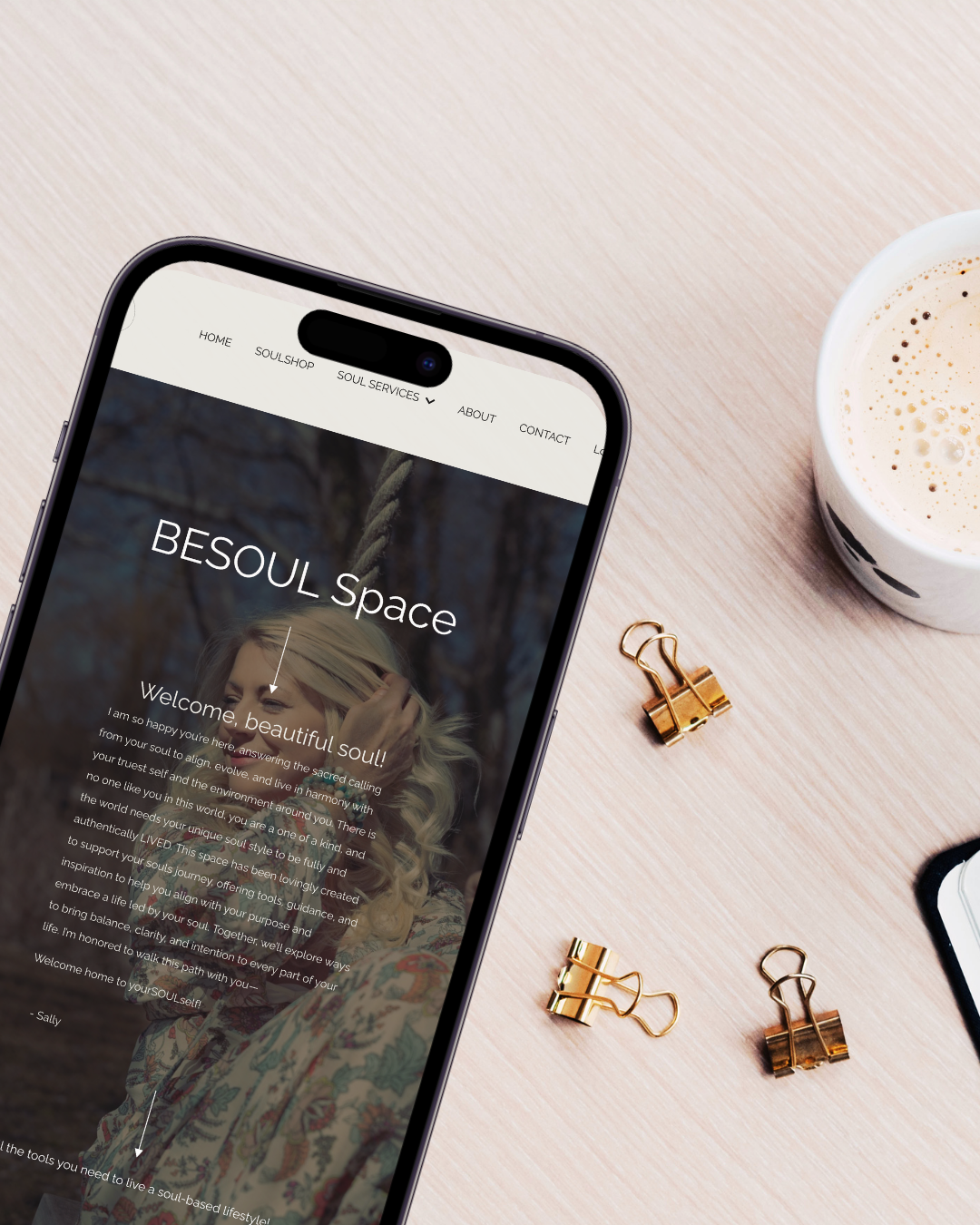 Smartphone displaying a website titled 'BESoul Space' with a welcome message, placed on a light wooden table. Next to the phone are gold binder clips and a cup of coffee.