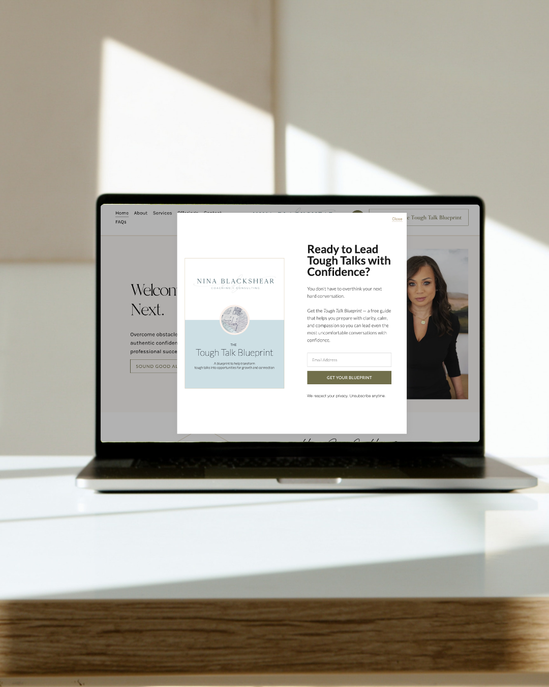 Laptop on a white desk with a light-colored wall and window in the background; the screen displays a pop-up for a coaching service called 'Tough Talk Blueprint' with a woman photograph and a sign-up form.