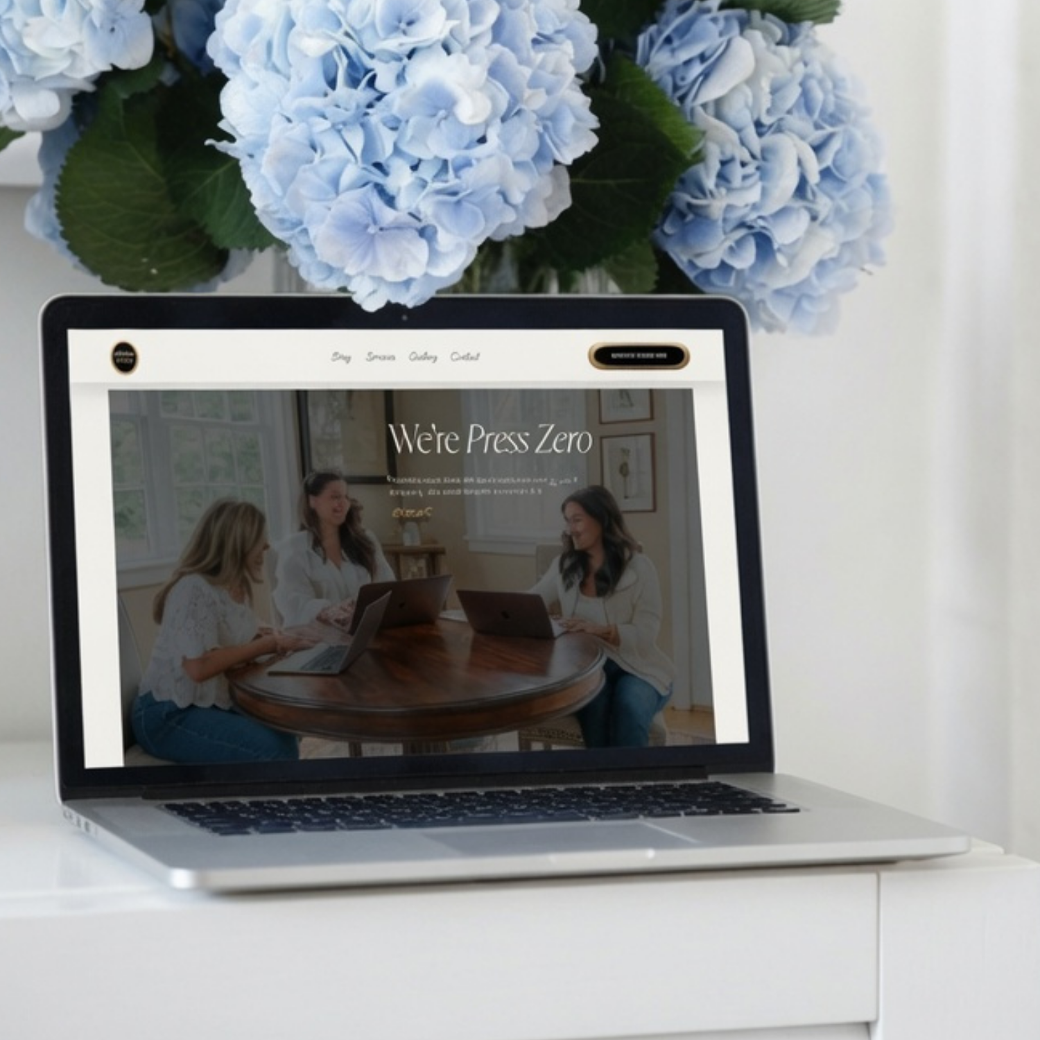 A laptop displaying a webpage with three women sitting at a table, working on laptops, in a bright room with framed pictures on the wall.