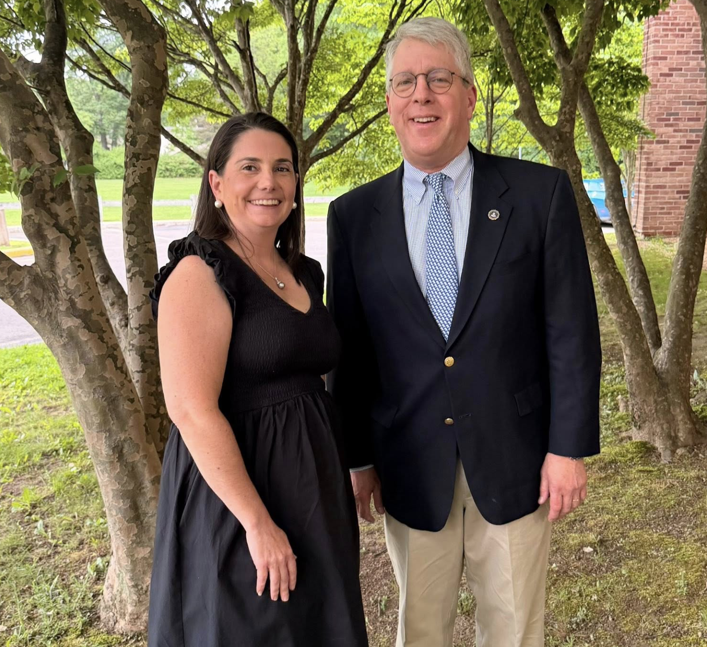 A woman and a man standing outdoors near a tree, smiling at the camera, dressed in formal attire, with green foliage and a brick building in the background.