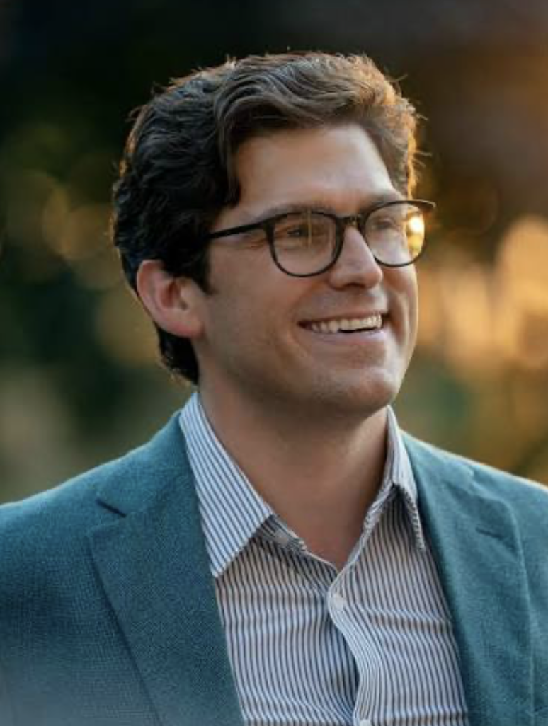 A smiling man with glasses wearing a teal blazer and a striped shirt outdoors during sunset.
