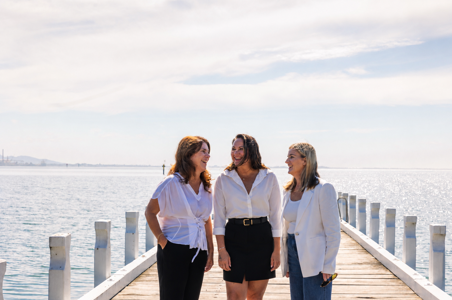 Three women standing on a wooden pier by the water, smiling and talking to each other on a sunny day.