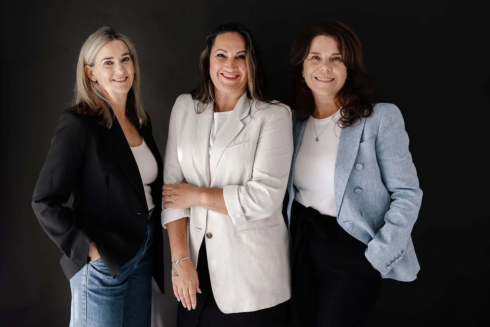 Three women standing together against a black background, dressed in business casual attire, smiling at the camera.