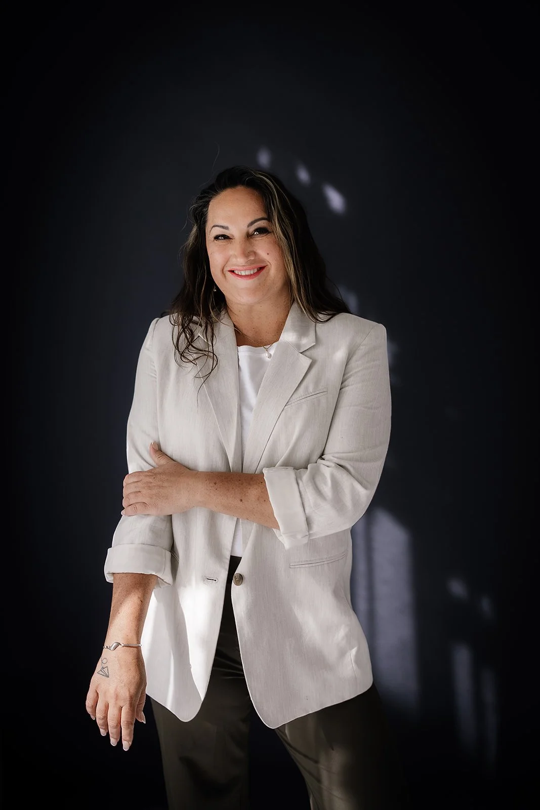 A woman smiling, wearing a beige blazer and a white shirt, standing against a black background.