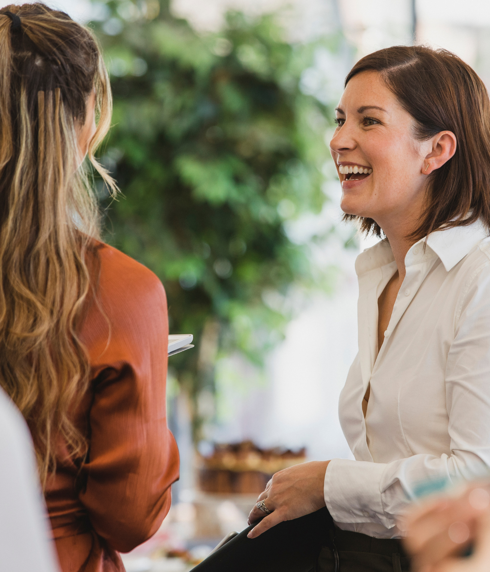 Two women engaged in a lively conversation in a bright, indoor setting with a blurred background of greenery.