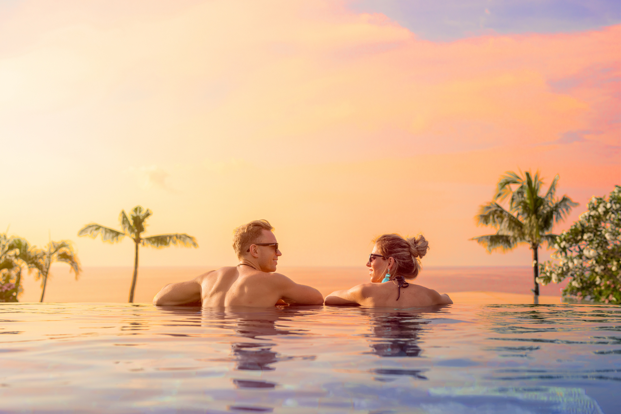 A couple relaxing in a swimming pool at sunset with palm trees and a colorful sky in the background.