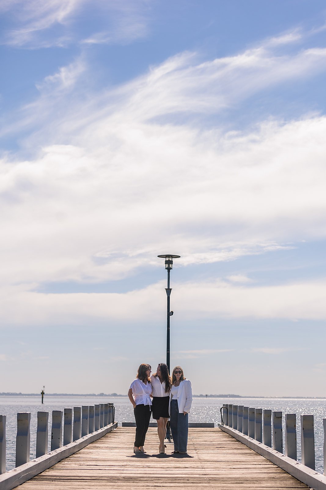 Three women in business casual clothing standing on a wooden pier by the water, with a cloudy sky above. They are talking and smiling, with a lamppost behind them.