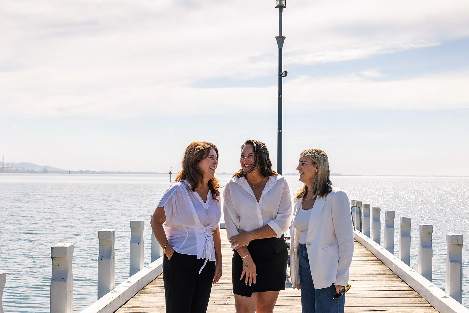 Three women are standing on a wooden dock by the water, smiling and talking to each other, with a lamppost behind them and a distant shoreline across the water.