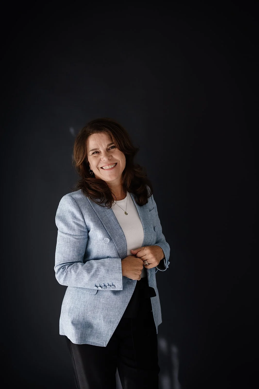 A smiling woman with brown hair wearing a light grey blazer over a white top, standing against a black background.