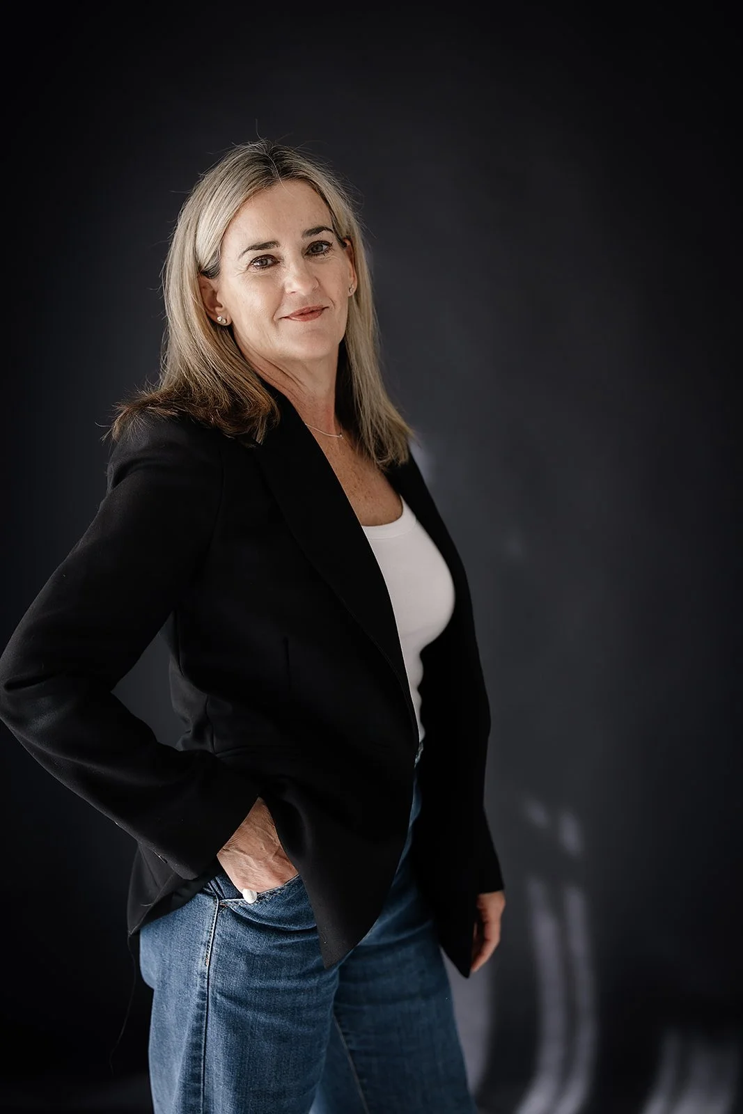 A woman with shoulder-length blonde hair wearing a black blazer, white top, and blue jeans standing with one hand in her pocket against a dark background.