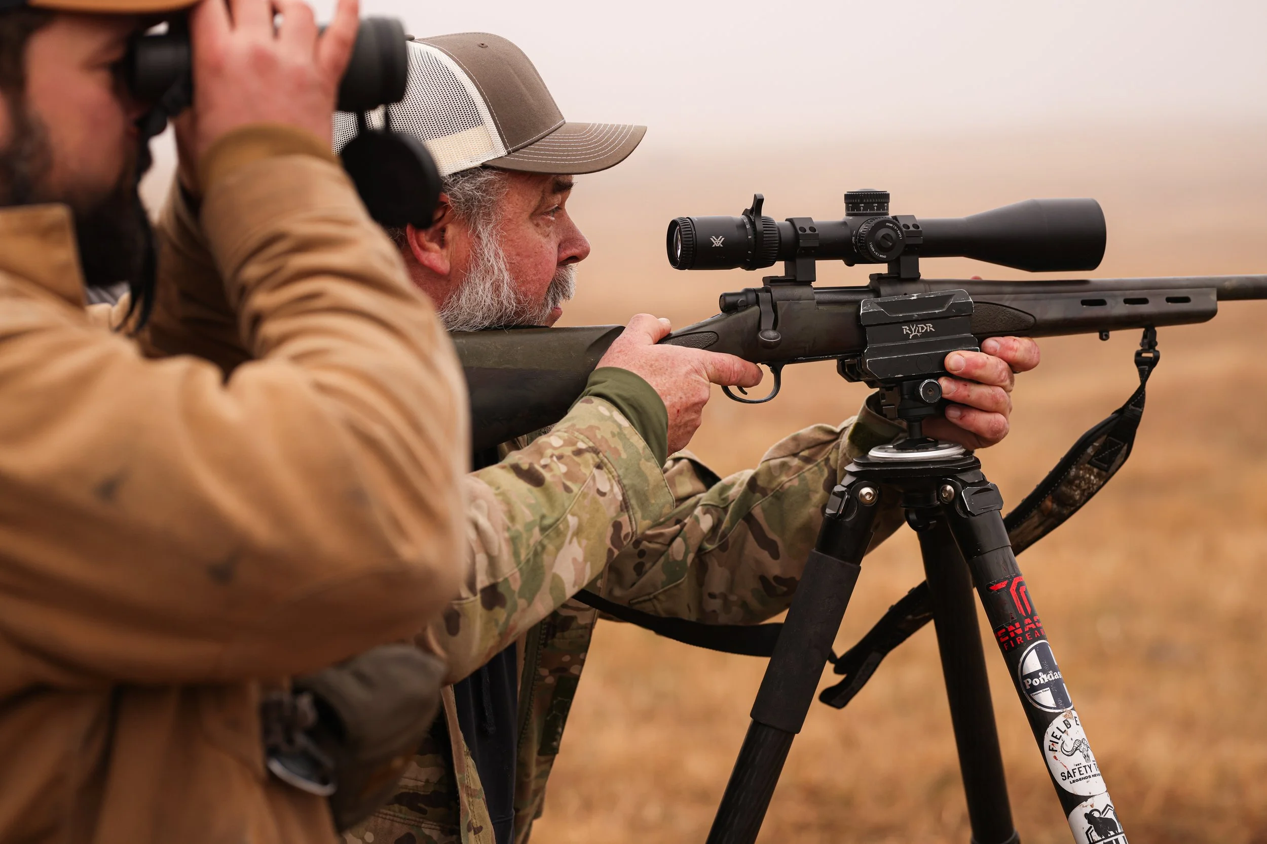 A man with gray hair and a beard is aiming through a rifle scope, using a spotting scope on a tripod, while another man watches with binoculars in a field.