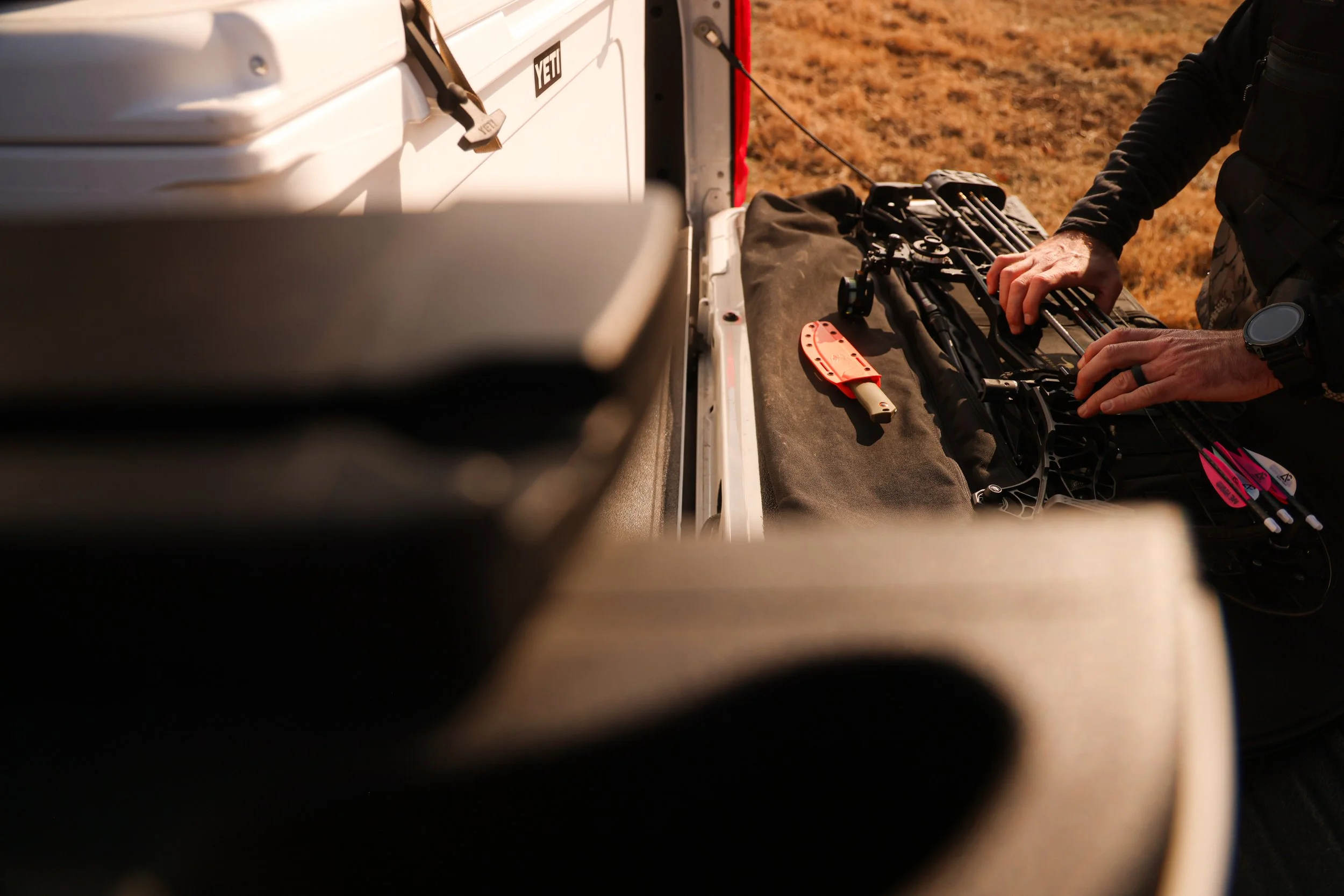 Person packing a compound bow into a soft case on the back of a vehicle outdoors.