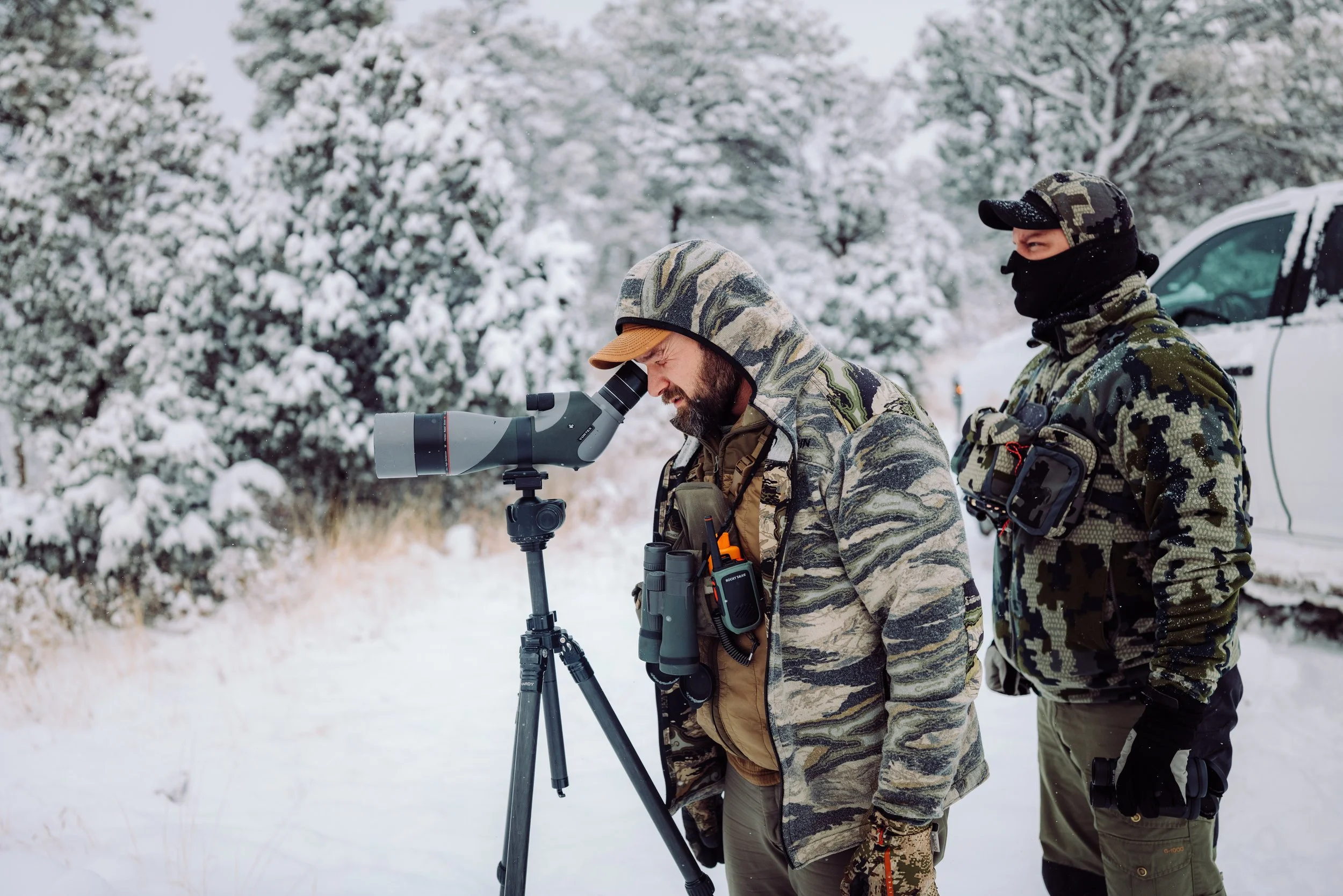 Two men dressed in camouflage and winter gear, looking through a telescope in a snowy outdoor setting with snow-covered trees and a vehicle in the background.