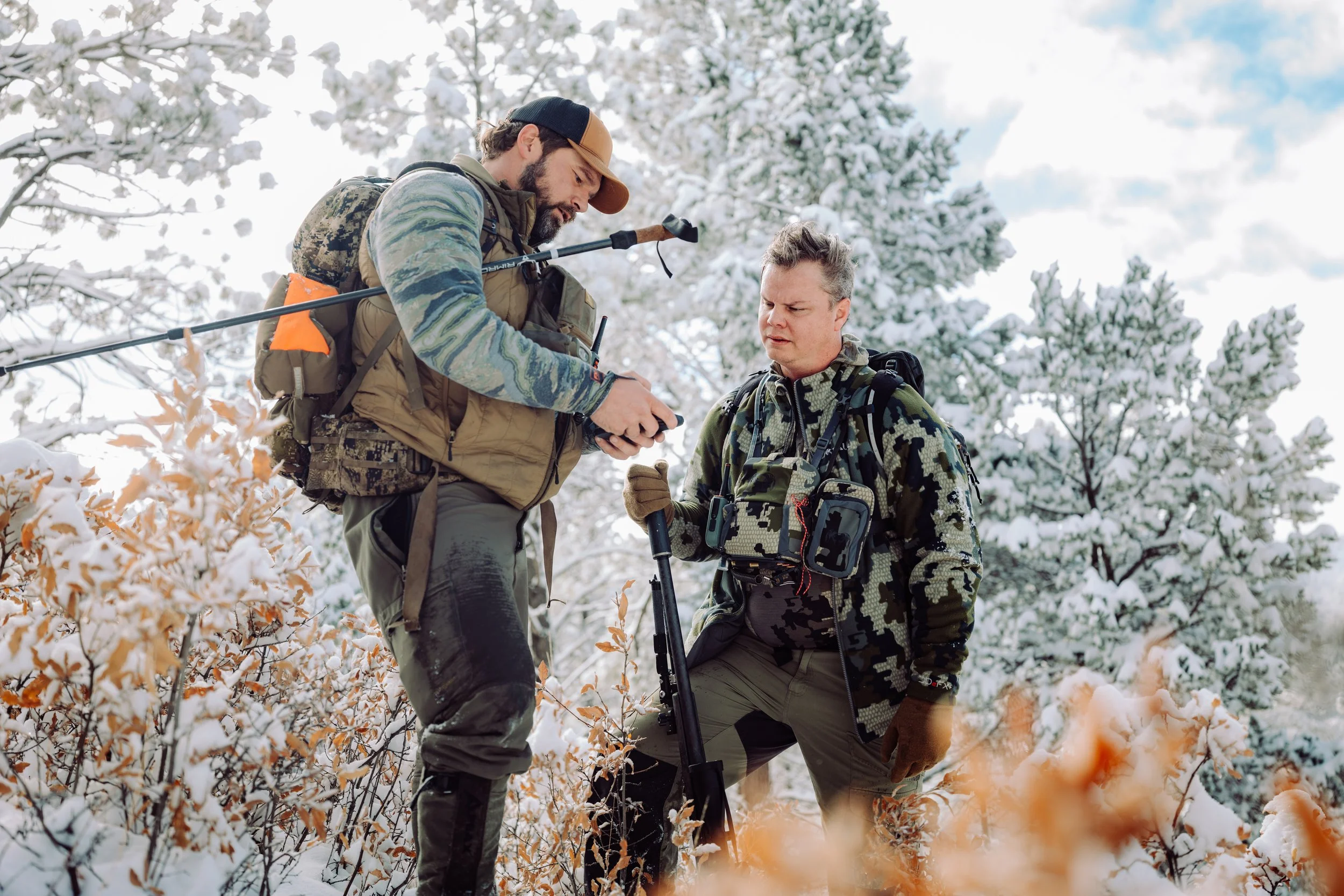 Two men in camouflage and outdoor gear standing in snowy forest, one showing something on a device to the other.