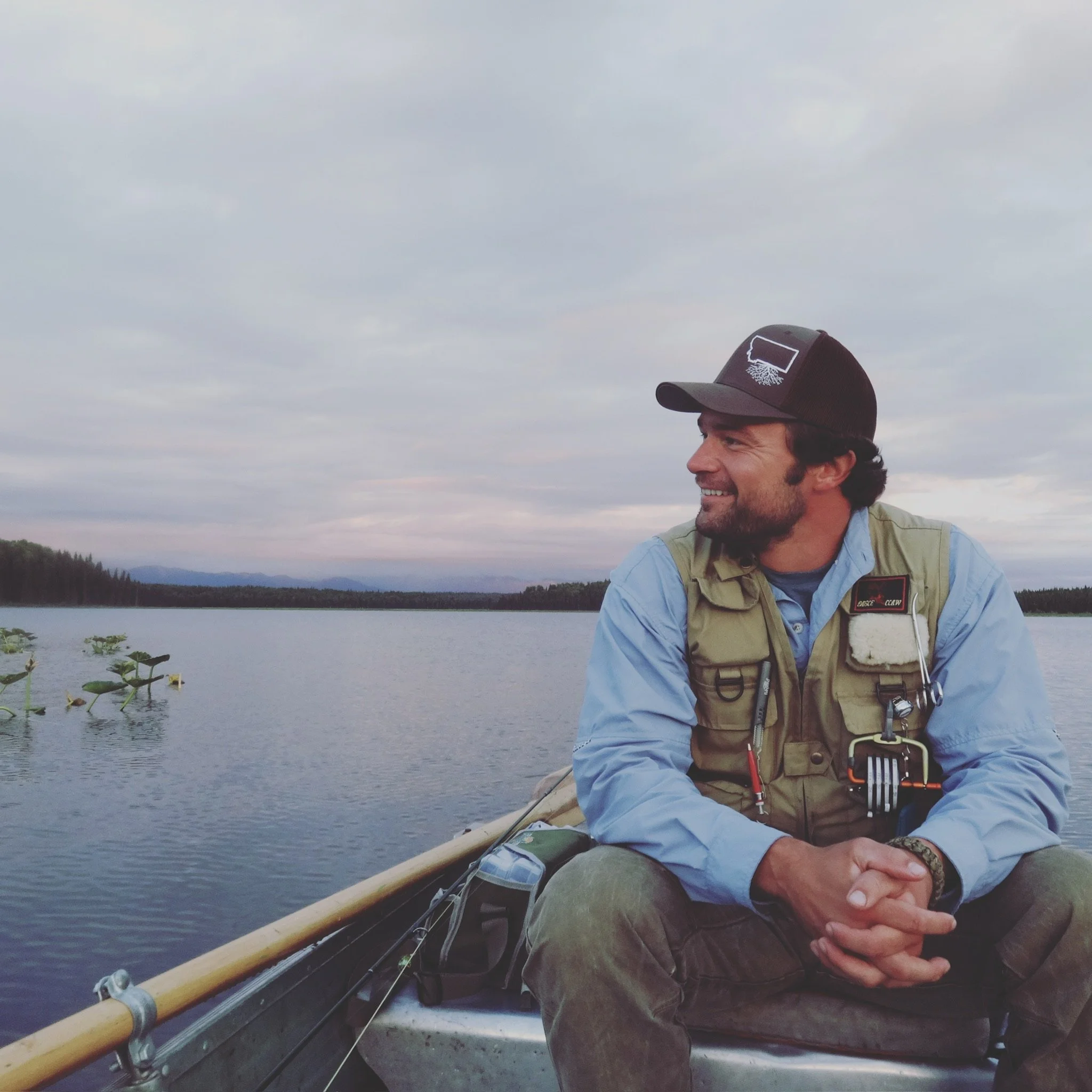 A man sitting in a boat on a lake, looking to his right, wearing a cap and vest, during daytime with cloudy sky.